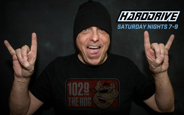 Man in a black beanie flashing rock-hand signs with both hands; wearing a 1029 FM The Hog T‑shirt, HardDrive graphic in the corner.