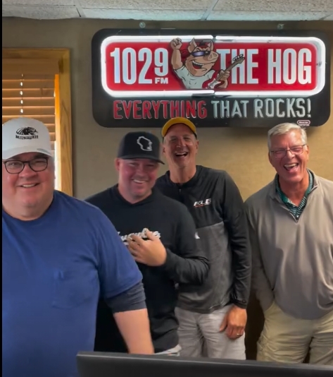 Four men posing in a radio studio under a sign that reads '1029 FM The Hog' and 'Everything that rocks!'