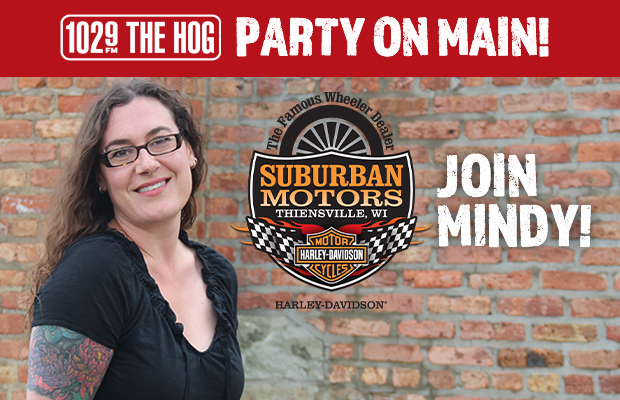 Smiling woman with glasses and tattooed arm stands in front of a brick wall with a Suburban Motors logo and 'Join Mindy!' for 102.9 The Hog.